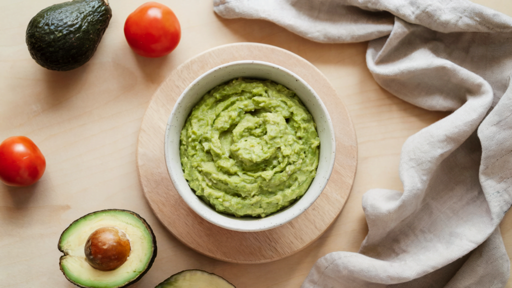 Gezonde avocado dip: guacamole! in een kom op een lichte houten ondergrond, omringd door ingrediënten zoals tomaten en avocados.